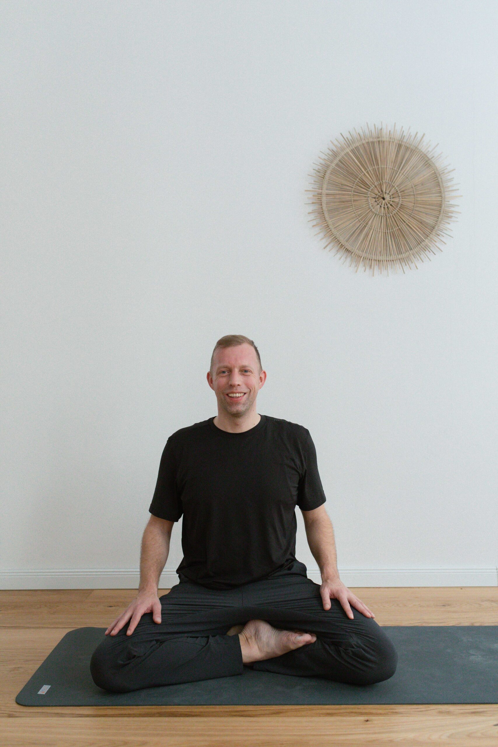 Chris sitzt lächelnd im Schneidersitz auf der Yogamatte